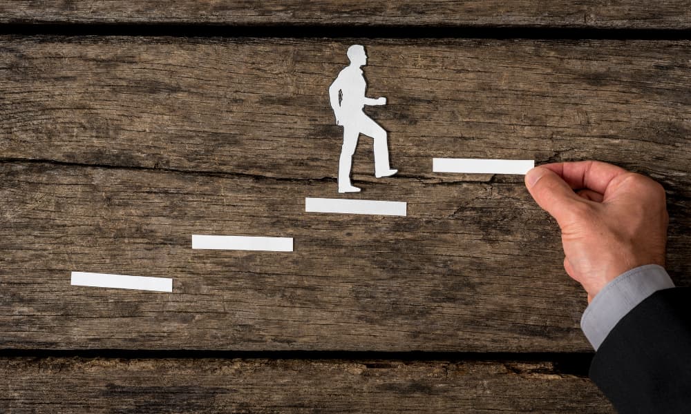 A paper figure ascending steps while a helpful hand adds the next platform.