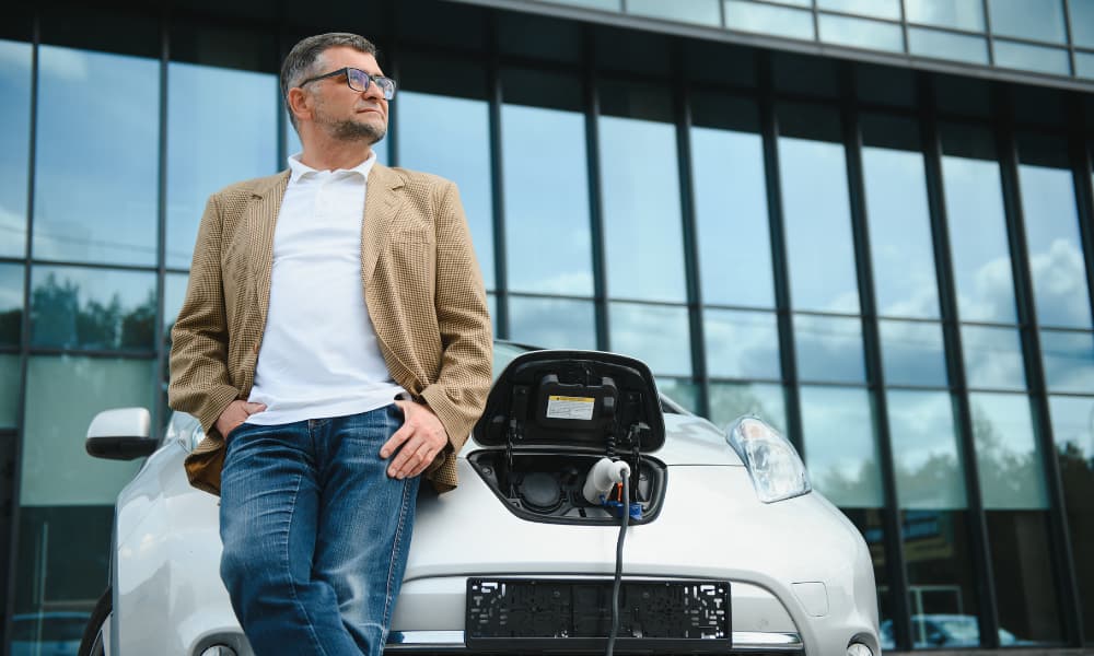 A casually dressed man leaning against an electric vehicle while it charges.