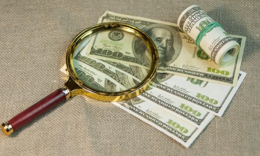 A collection of $100 bills laid out on beige fabric under a brass magnifying glass with a burgundy handle.