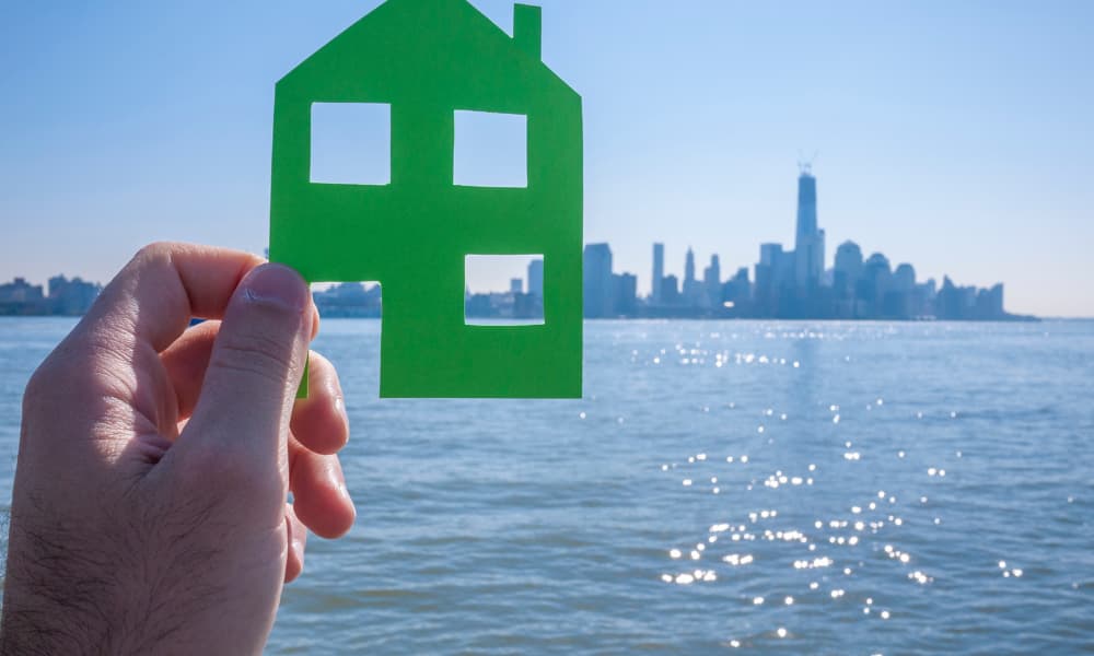 A hand is holding a green paper cutout of a house against the sunny New York city skyline viewed from across the water.